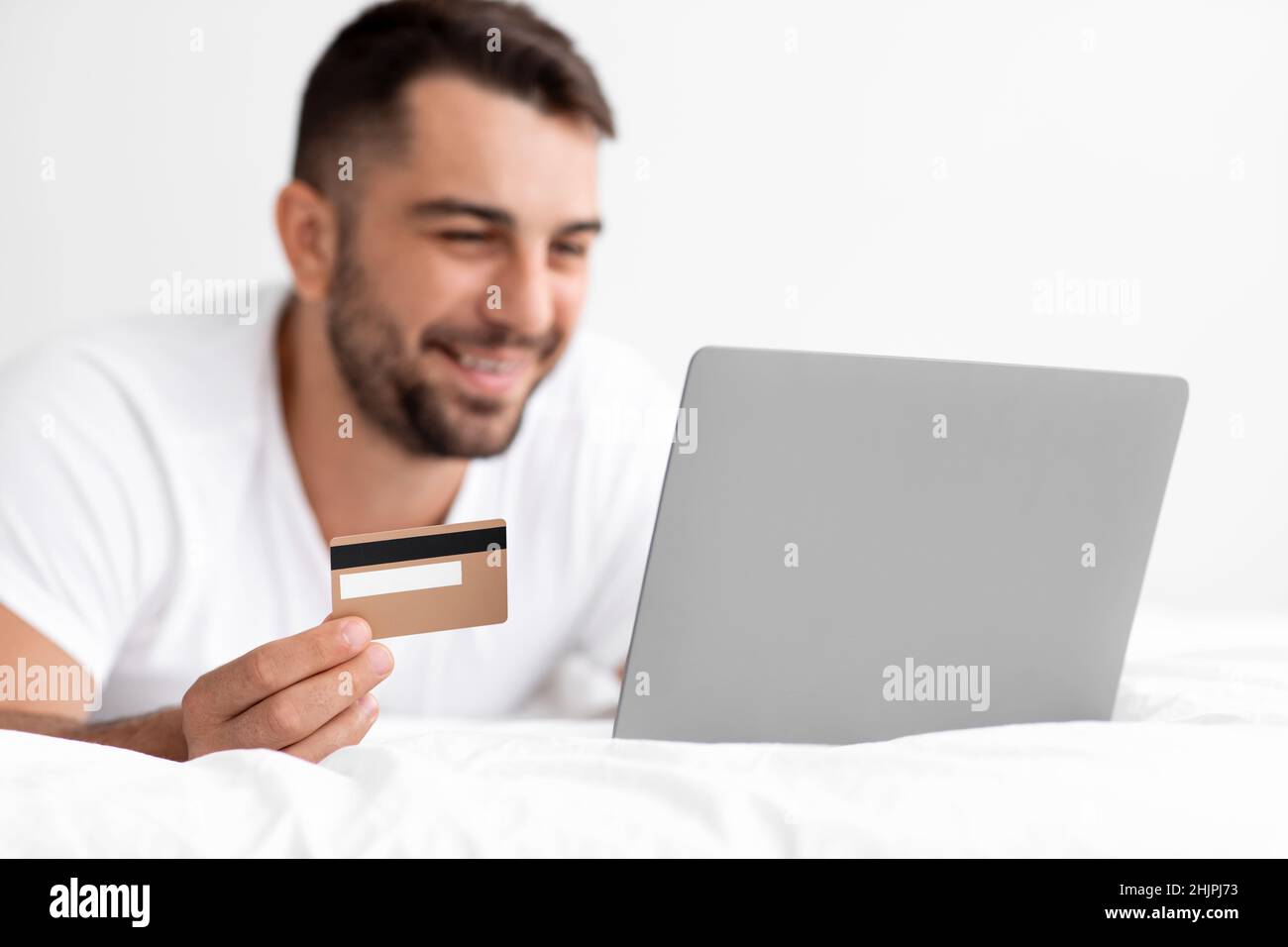 Felice caucasico attraente maschio millenario in bianco t-shirt acquistare online, ordini di acquisto su pc e mostra carta di credito Foto Stock