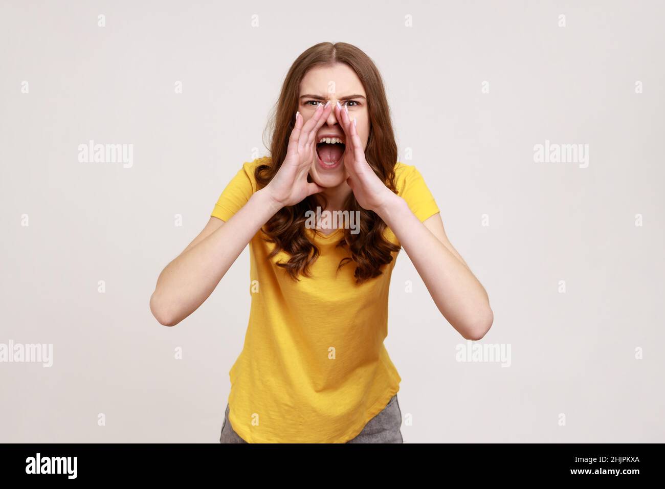 Ritratto di ragazza adolescente irritata furiosa con capelli ondulati marroni in maglietta casual gialla urlando forte, sperimentando forte aggressività. Studio interno girato isolato su sfondo grigio. Foto Stock