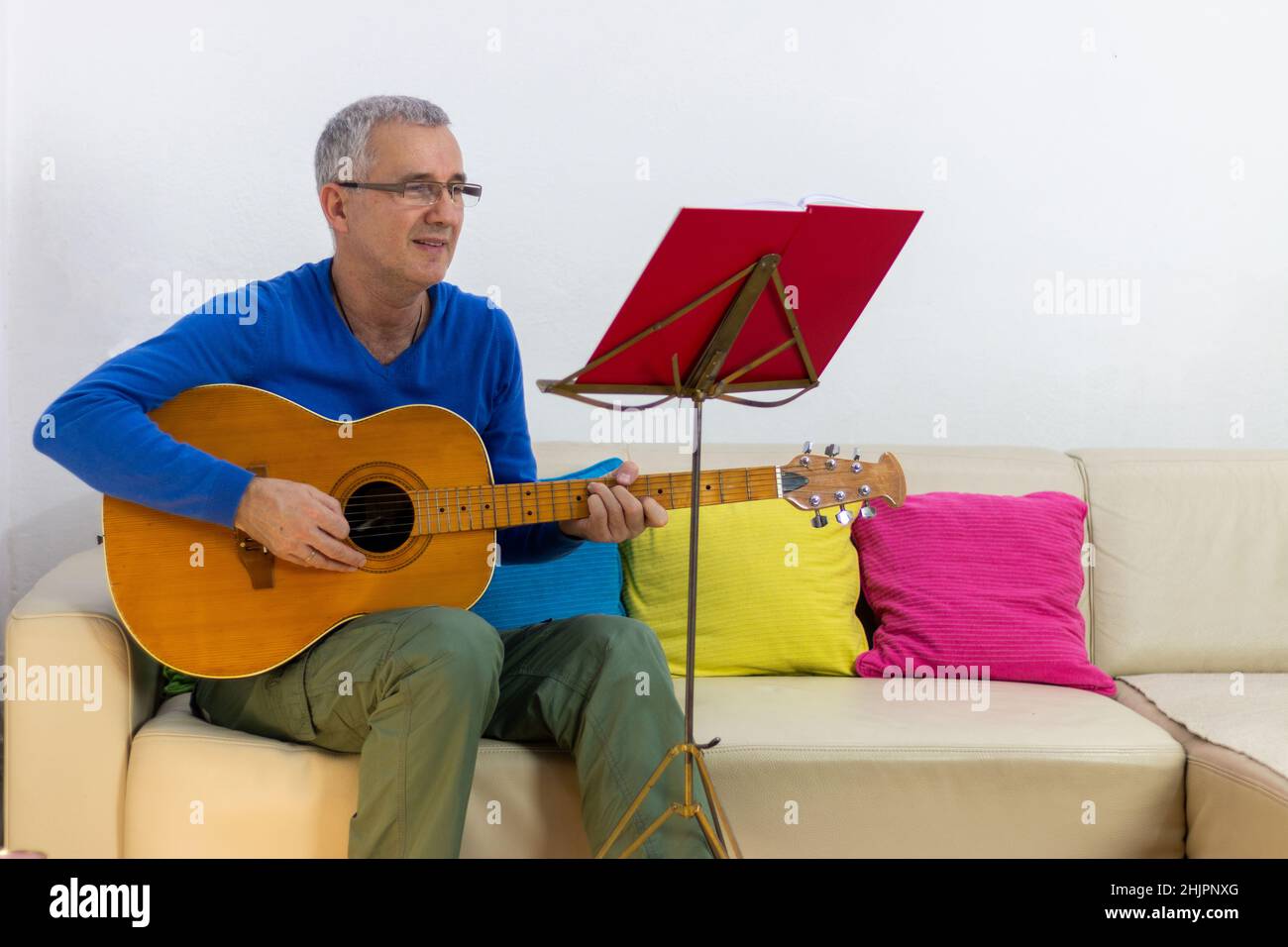 Buon osservare uomo grigio maturo dei capelli esercitandosi a suonare la chitarra seduta sul divano nel paese Foto Stock