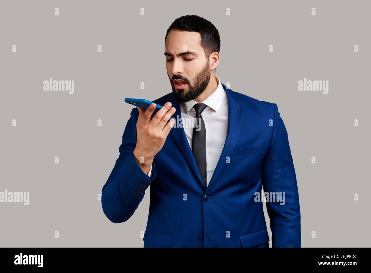 Uomo portatore che tiene il telefono cellulare, impartendo comandi all'assistente vocale, lasciando un messaggio vocale o registrando note audio, indossando un abito di stile ufficiale. Studio interno girato isolato su sfondo grigio. Foto Stock