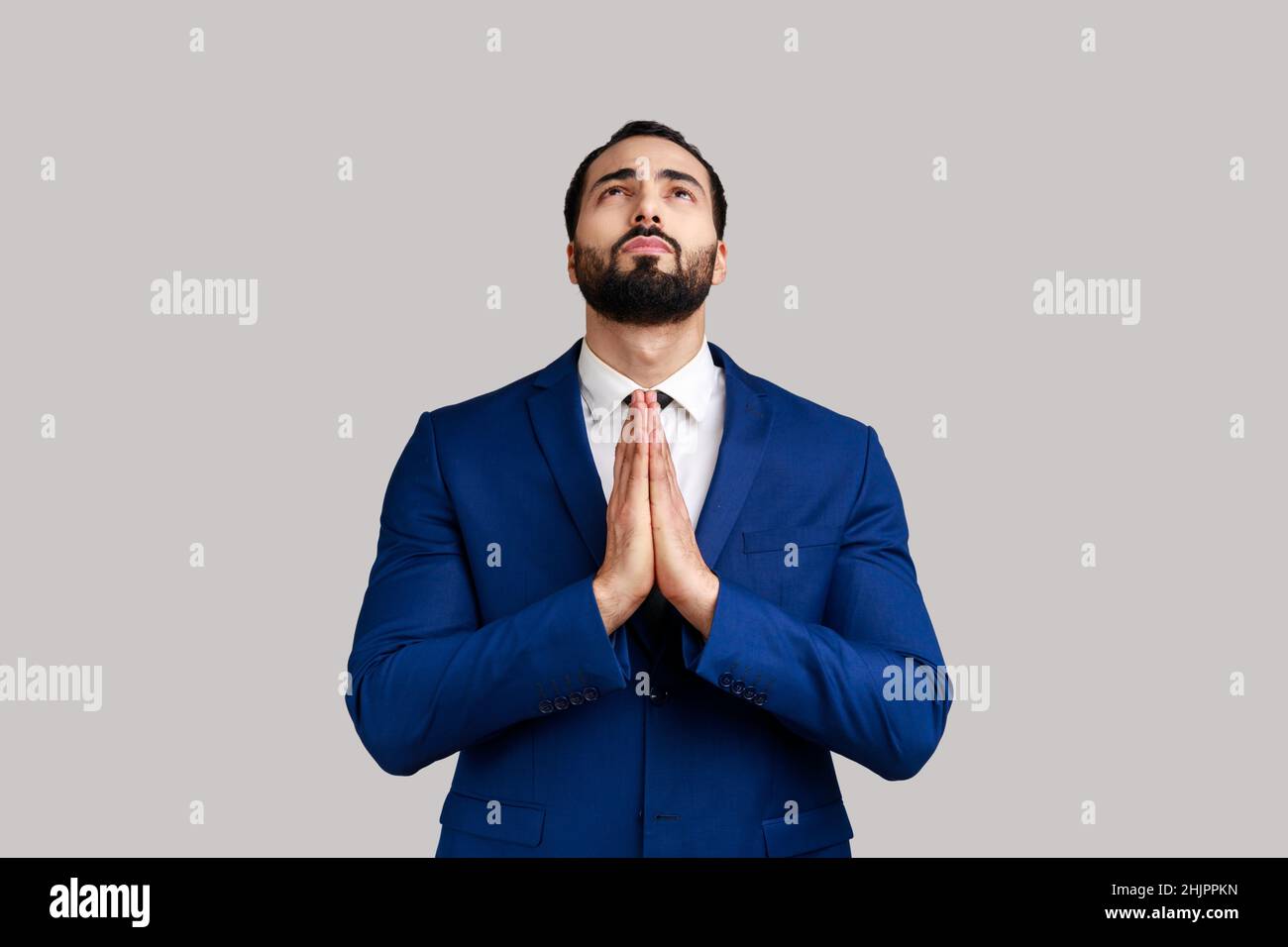 Ritratto di bel barbuto uomo che prega di cuore, sentendosi colpevole, supplicando il cielo implorando aiuto, indossando vestito di stile ufficiale. Studio interno girato isolato su sfondo grigio. Foto Stock