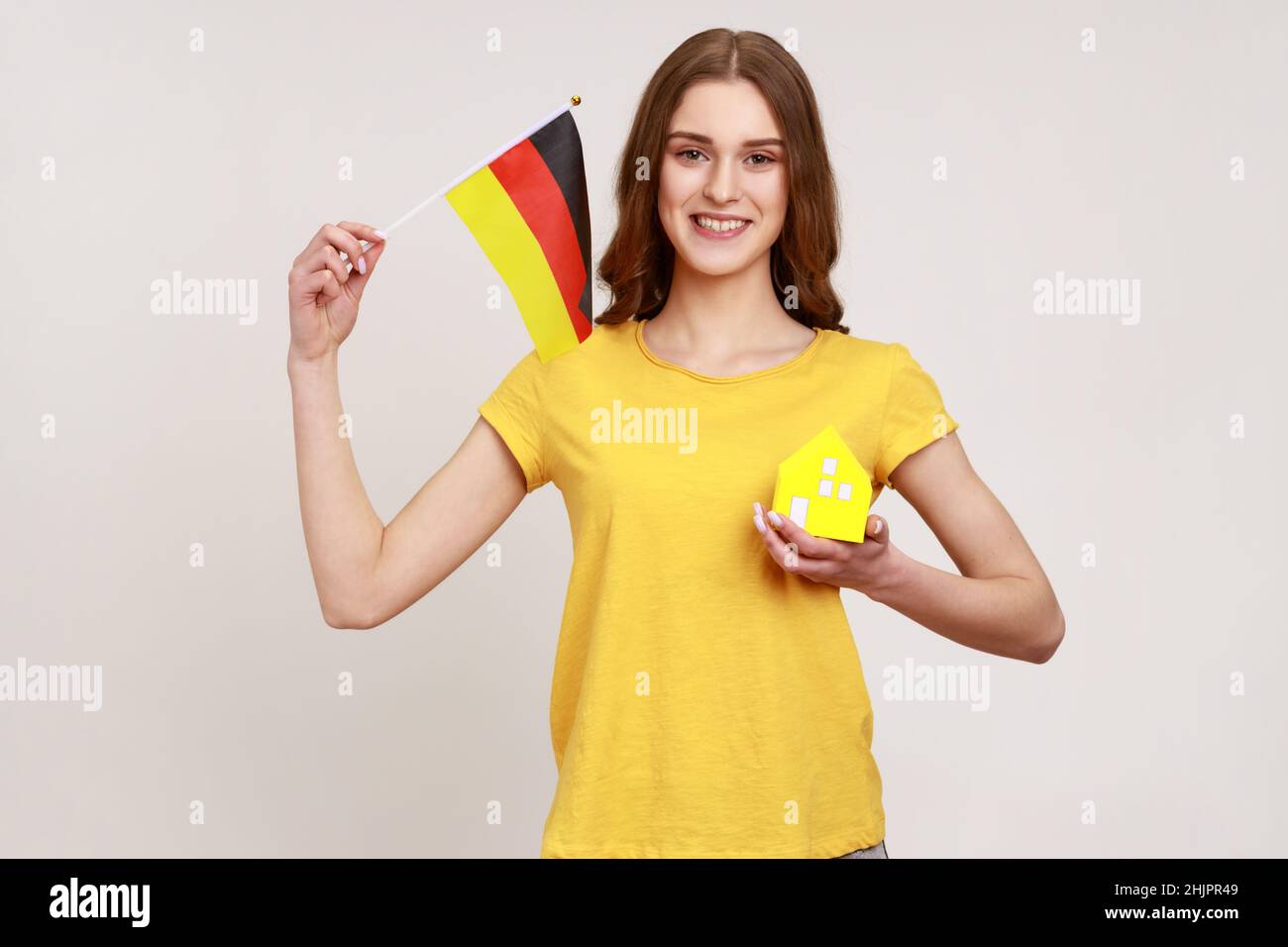 Ragazza sorridente positiva in giallo casual T-shirt mostrando piccola casa di carta e tenendo bandiera della germania, soddisfatto con l'acquisto di alloggio. Studio interno girato isolato su sfondo grigio. Foto Stock