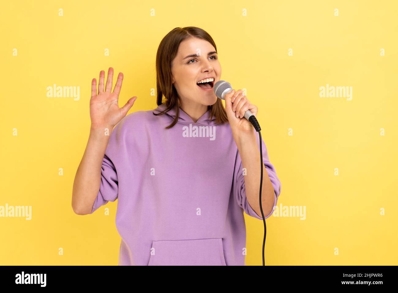 Felice positiva giovane donna in piedi con microfono cantare canzoni in karaoke con braccio sollevato, divertirsi da solo, indossando felpa con cappuccio viola. Studio interno girato isolato su sfondo giallo. Foto Stock