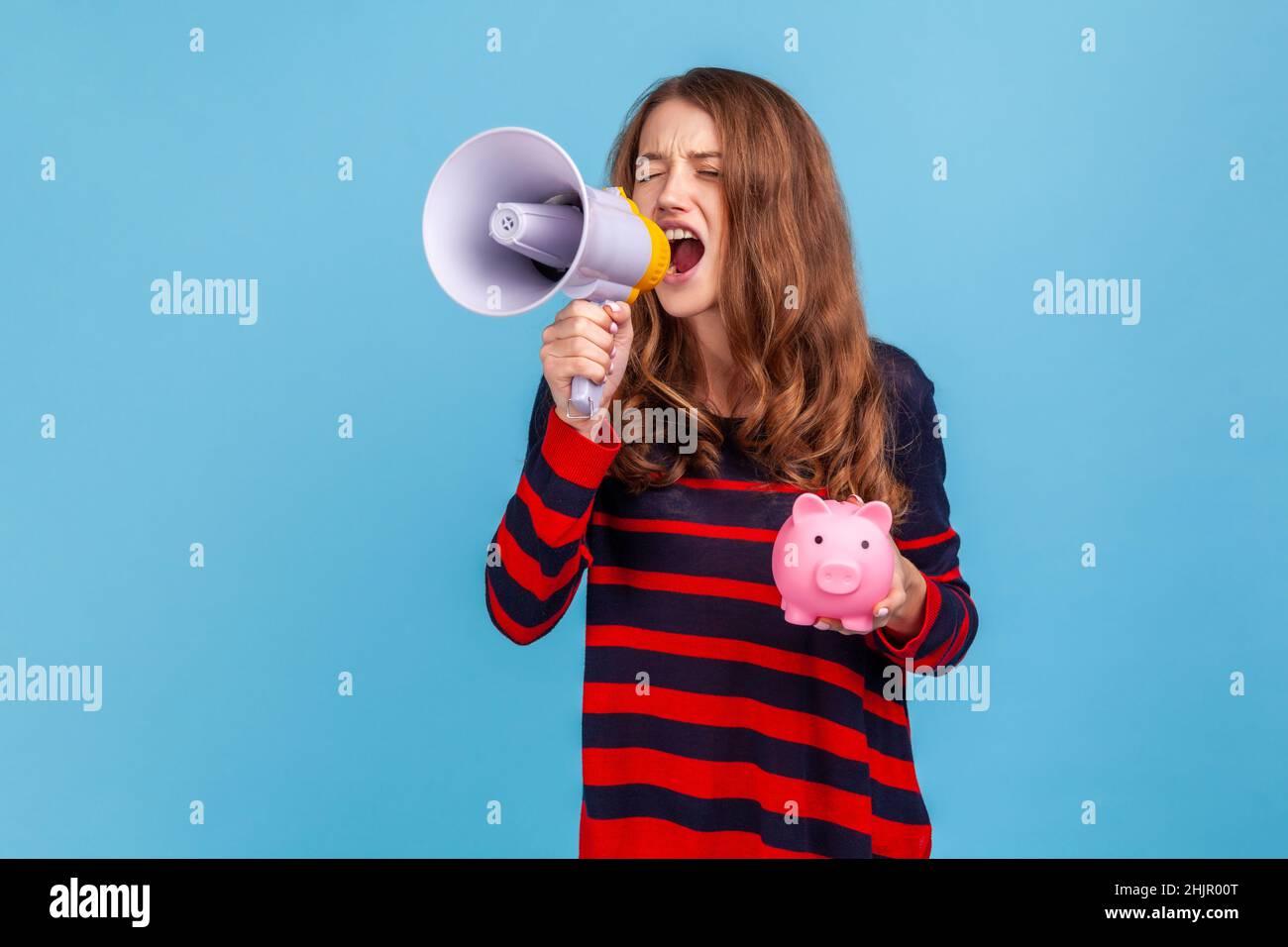 Ritratto di donna indossando a righe casual stile maglione, tenendo Piggy banca e urla in megaphone, annunciando offerte speciali banca. Studio interno girato isolato su sfondo blu. Foto Stock