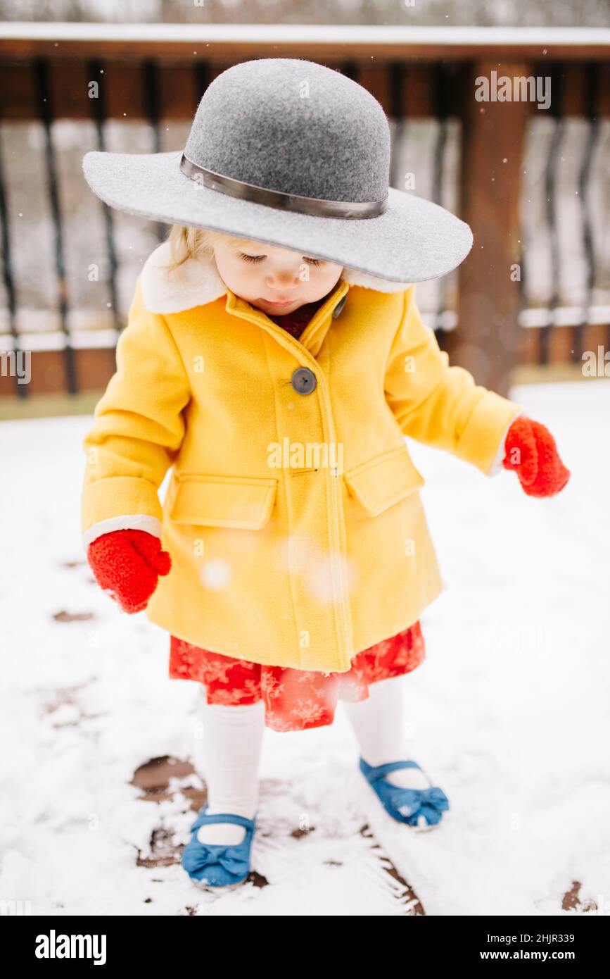 Bambina in piedi nella neve con cappotto giallo, cappello e guanti rossi Foto Stock