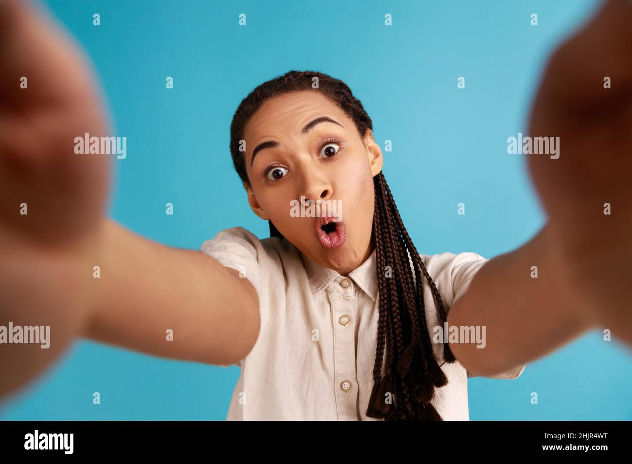 Donna stupita con dreadlocks nero ha sorpreso l'espressione del volto, allunga il braccio per fare selfie, esprime emozioni sincere, punto di vista foto. Studio interno girato isolato su sfondo blu. Foto Stock