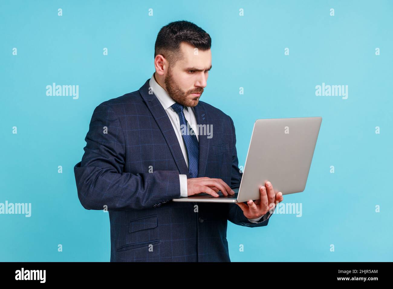 Giovane adulto concentrato vestito ufficiale di stile che tiene il laptop nelle mani, lavorando sul computer, guardando il display con espressione seria. Studio interno girato isolato su sfondo blu. Foto Stock