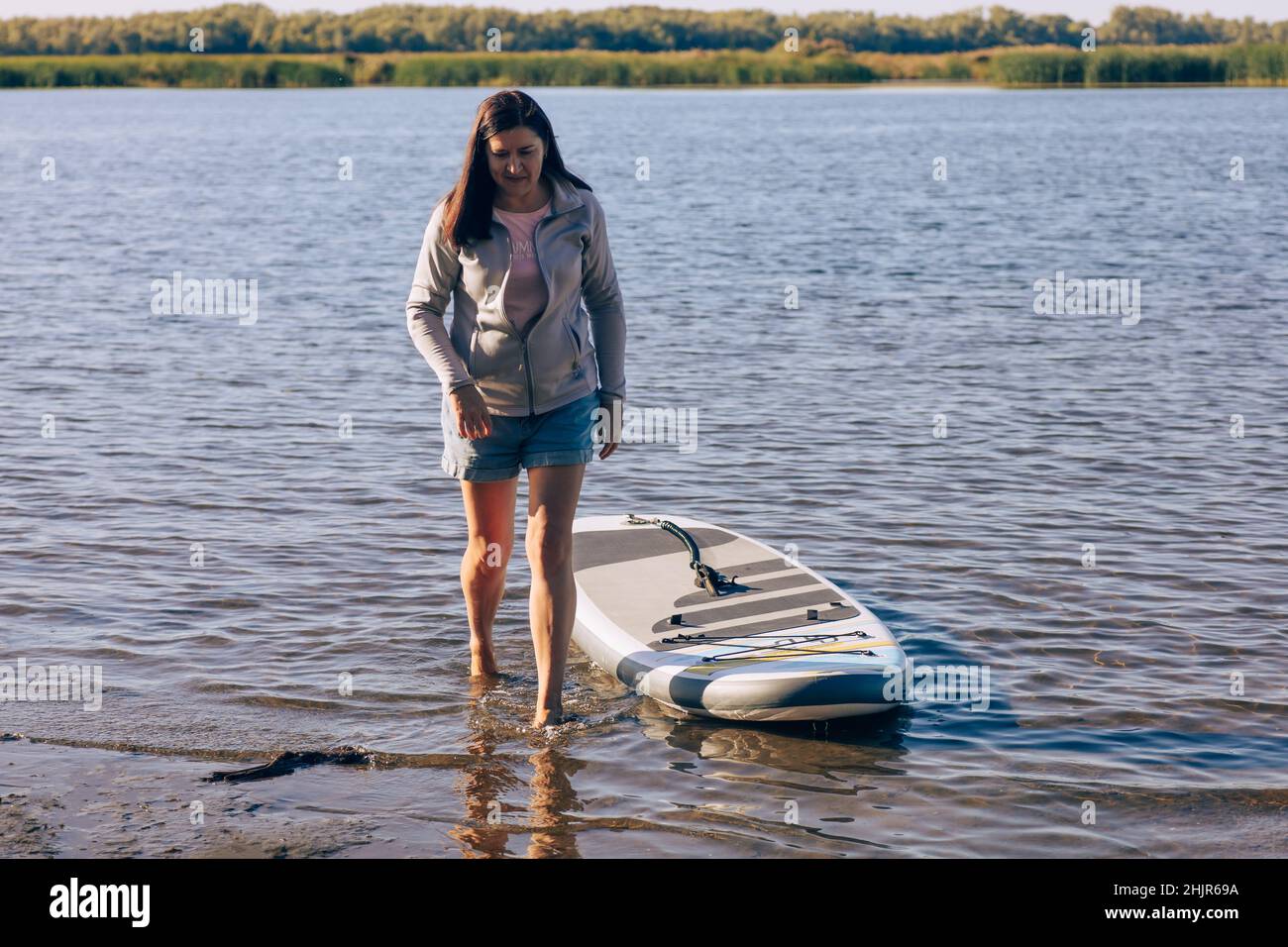 Donna stanca di mezza età che cammina fuori dall'acqua del lago con la tavola di sup dietro con canne e alberi sullo sfondo indossando camicie e maglione grigio. Attivo Foto Stock