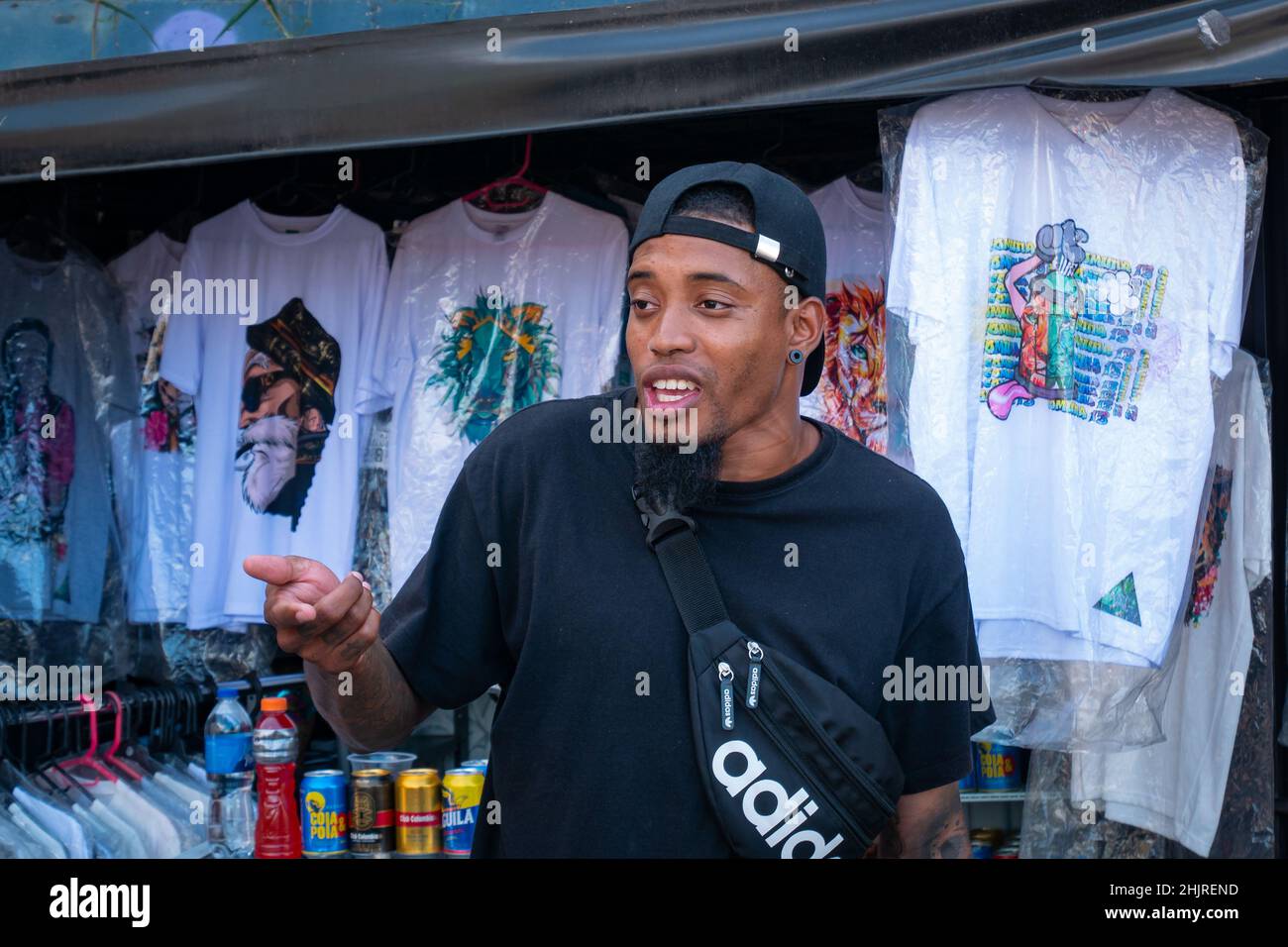 Medellin, Antioquia, Colombia - Novembre 11 2021: Giovane uomo latino con T-Shirt e cappellino nero sta vendendo vestiti e birre Foto Stock