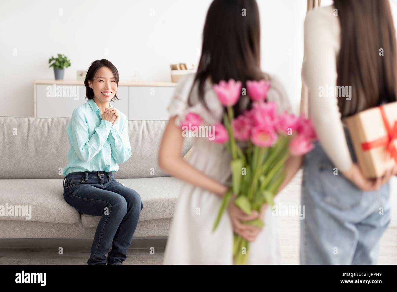 Piccola ragazza asiatica con la madre che nasconde il regalo e i fiori per la donna matura dietro le loro spalle a casa Foto Stock