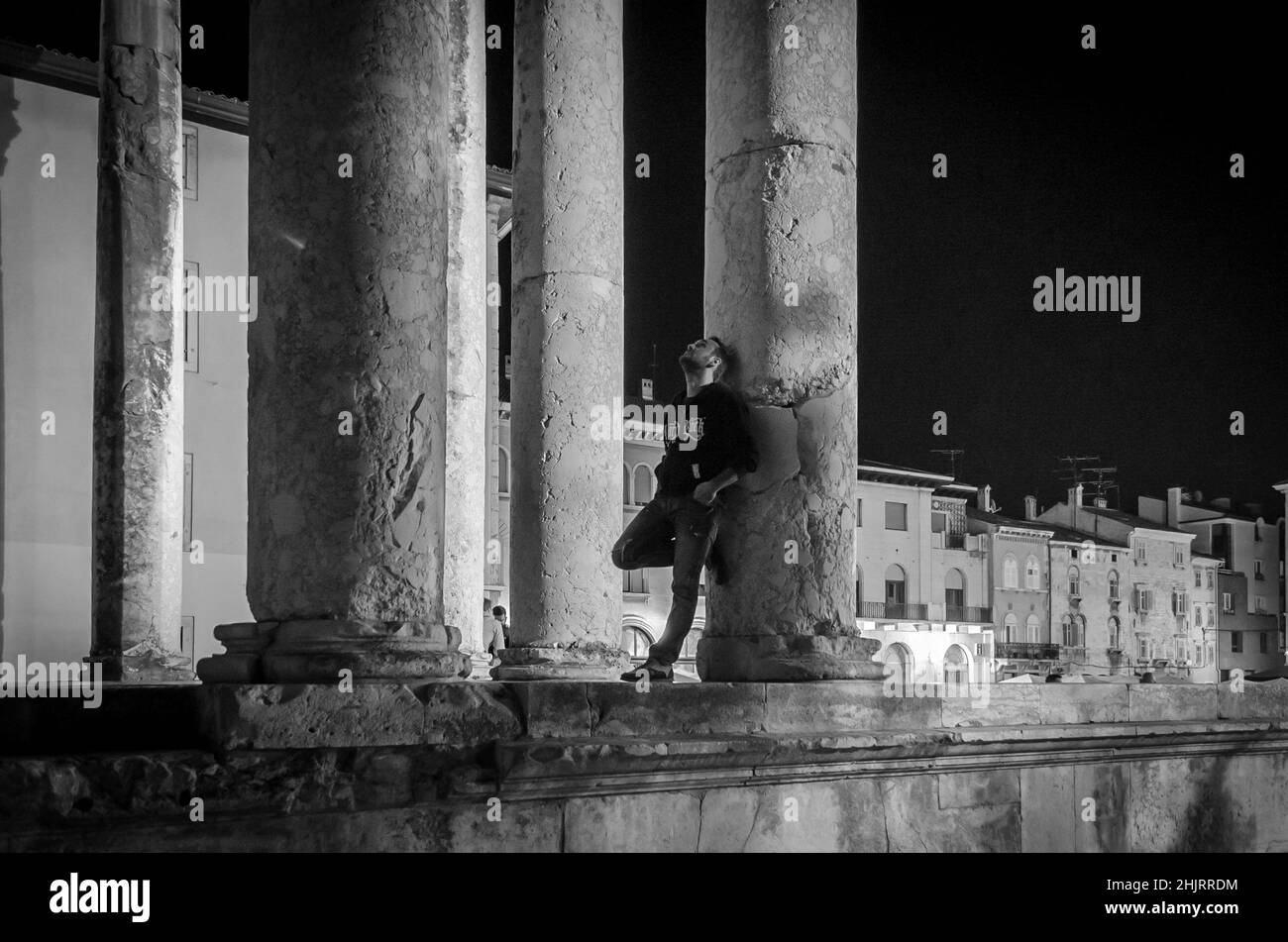 Modello maschile posa e posa sulla colonna dell'antico tempio romano di Augusto a Pula, Croazia, di notte. Fotografia in bianco e nero Foto Stock