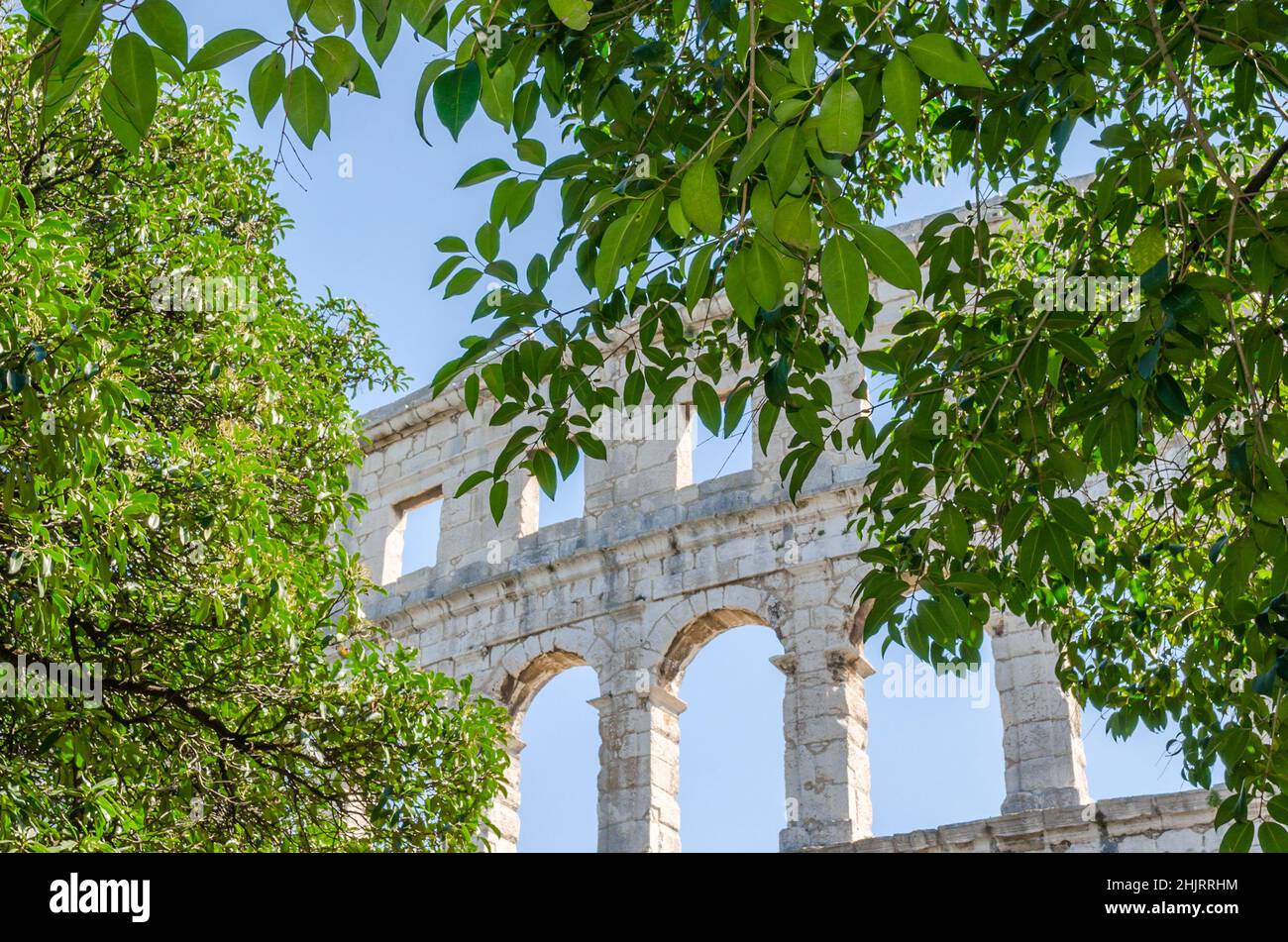 La Pula Arena attraverso grandi alberi con foglie verdi. Antico Anfiteatro romano con mura ad arco restaurate situato in Croazia. Ben conservato Monumento. Foto Stock