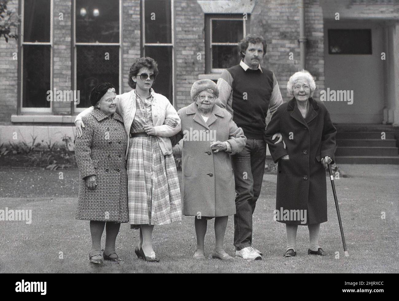 1980s, storica, tre donne anziane a ragion veduta nei loro cappotti che si ergono fuori nel giardino della loro casa del vecchio popolo, insieme con il marito e la squadra della moglie che si occupano della casa di cura, Engerd, Regno Unito. Foto Stock