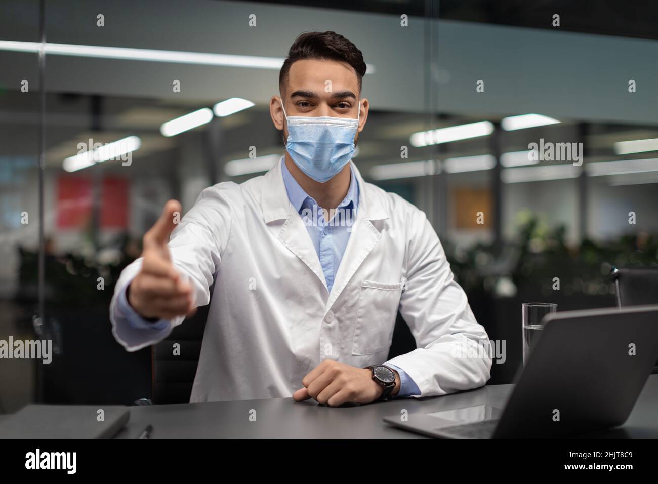 Medico in maschera seduta alla scrivania, saluto paziente Foto Stock
