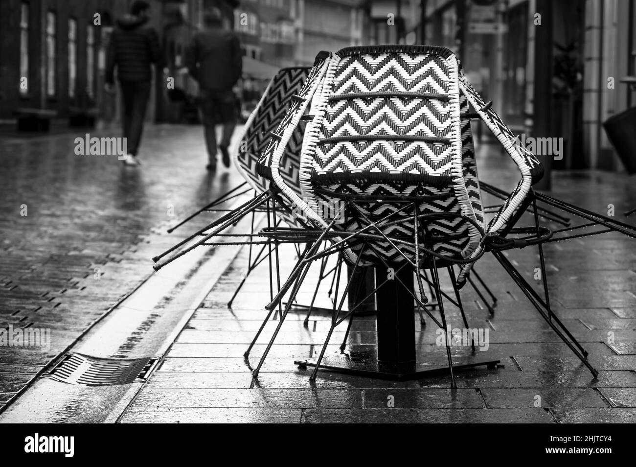 Sedie assemblate dal caffè marciapiede in una strada piovosa e pedoni da dietro passando vicino, immagine della vita cittadina in bianco e nero, spazio copia, selezionato Foto Stock