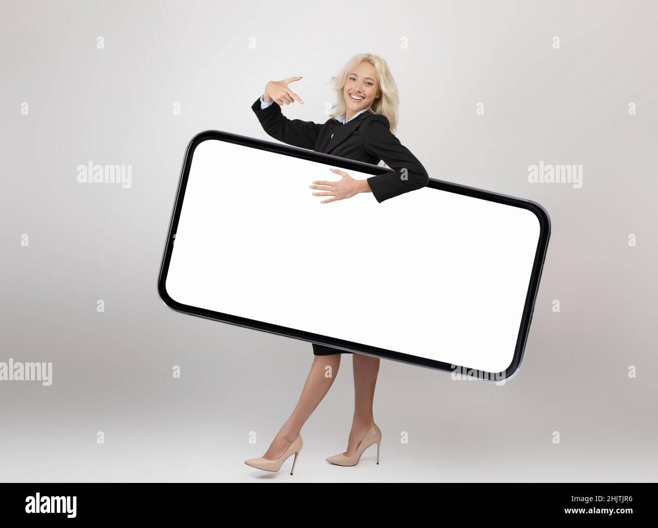 Scatto a tutta la lunghezza del corpo di una donna d'affari felice che trasporta un grande smartphone con schermo vuoto e punta allo schermo del gadget Foto Stock