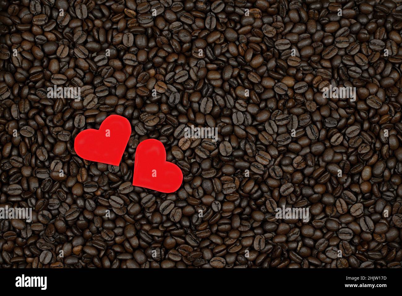 I chicchi di caffè di San Valentino sono sparsi sulla superficie e due cuori rossi brillano sul loro sfondo Foto Stock