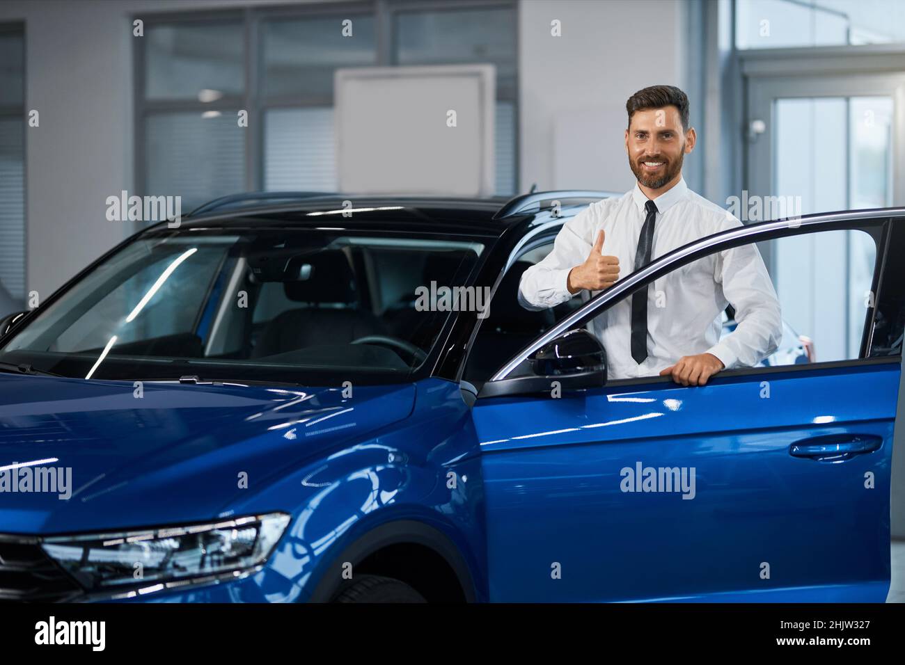 Uomo d'affari sorridente in abbigliamento formale in piedi vicino a un'auto blu di lusso e gesturando il pollice sulla fotocamera. Acquisto di nuovi mezzi di trasporto presso il salone auto. Foto Stock