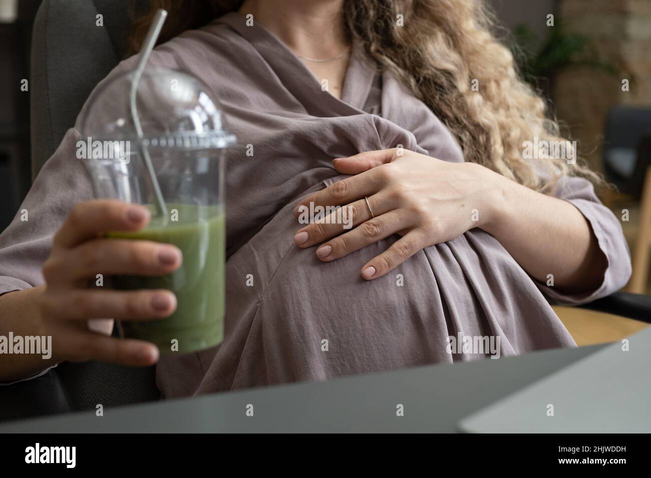 Giovane donna incinta in vestito grigio che tiene la mano sul ventre mentre si siede in poltrona dalla scrivania e ha un cocktail di frutta o un frullato Foto Stock