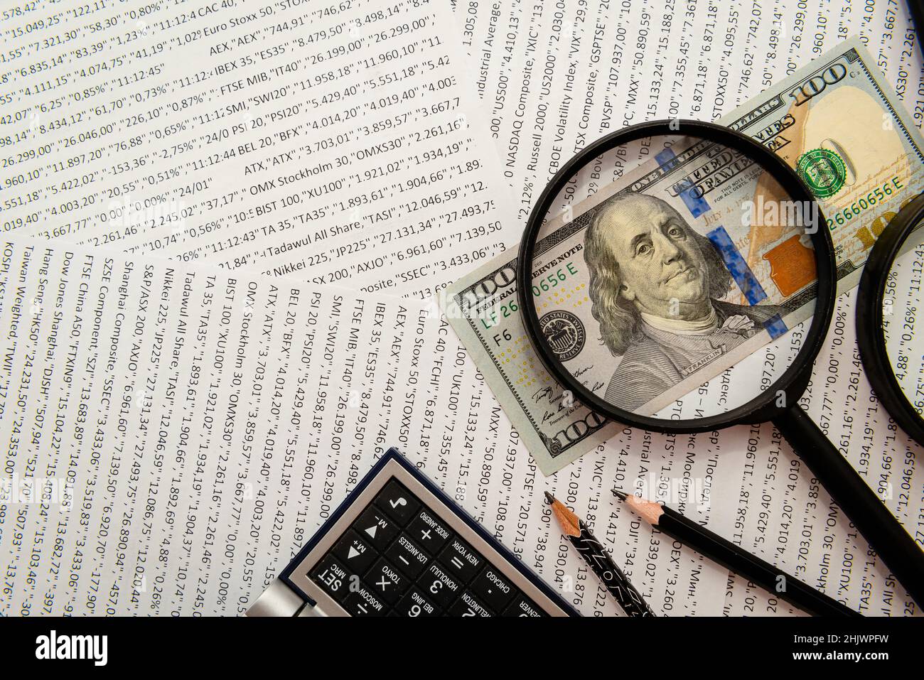 Una fattura del cento dollari sotto una lente d'ingrandimento contro lo sfondo delle tabelle e delle citazioni di azione. Affari e Finanza Foto Stock