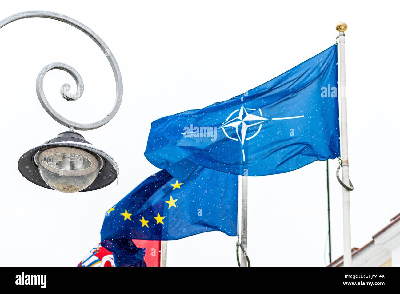 La bandiera della NATO e dell'Unione europea si è radunata in un cielo grigio in una fredda giornata invernale nevosa con un concetto di lampione, gas ed energia Foto Stock