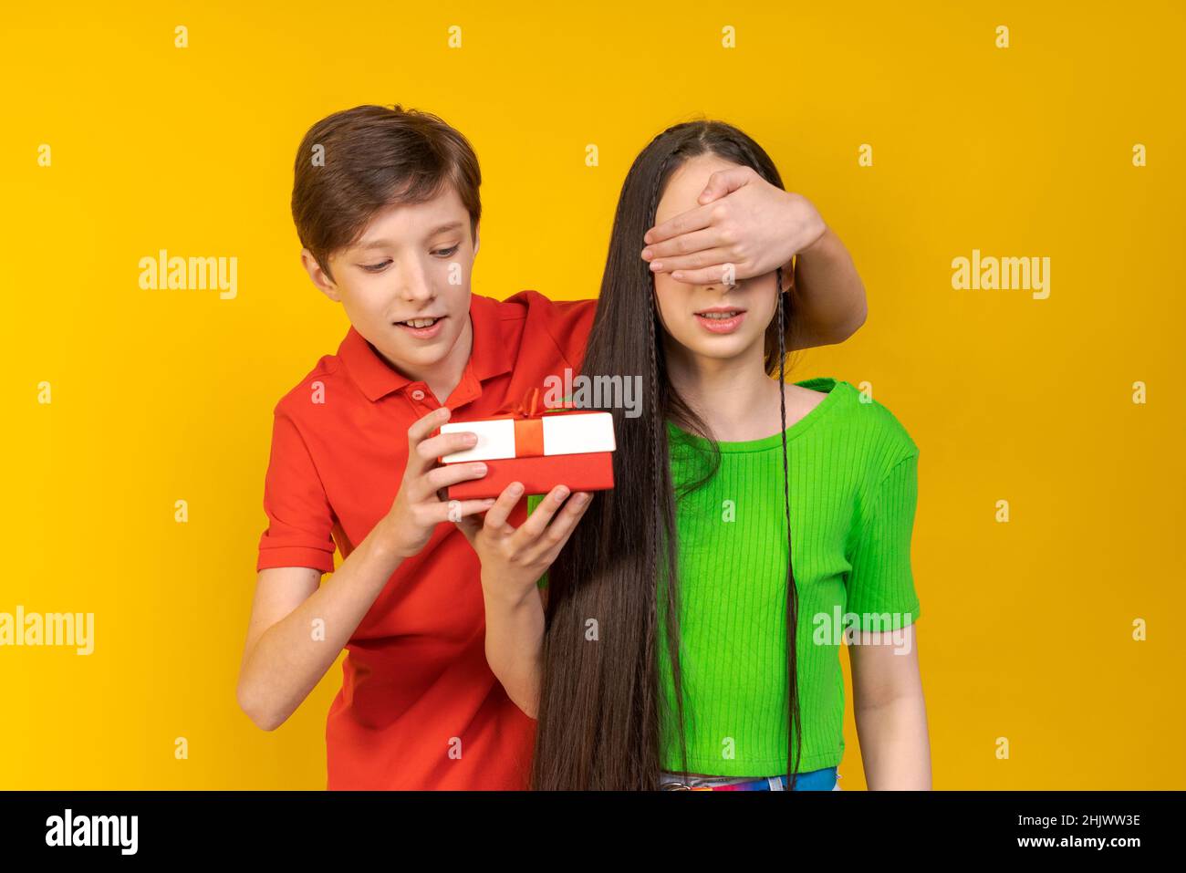 Chi è questo. Uomo caucasico positivo che copre gli occhi della ragazza dalla parte posteriore mentre sta dietro di lei e sorridente, adolescenti felici che si divertono sopra sfondo giallo studio, indossando abiti casual colore Foto Stock