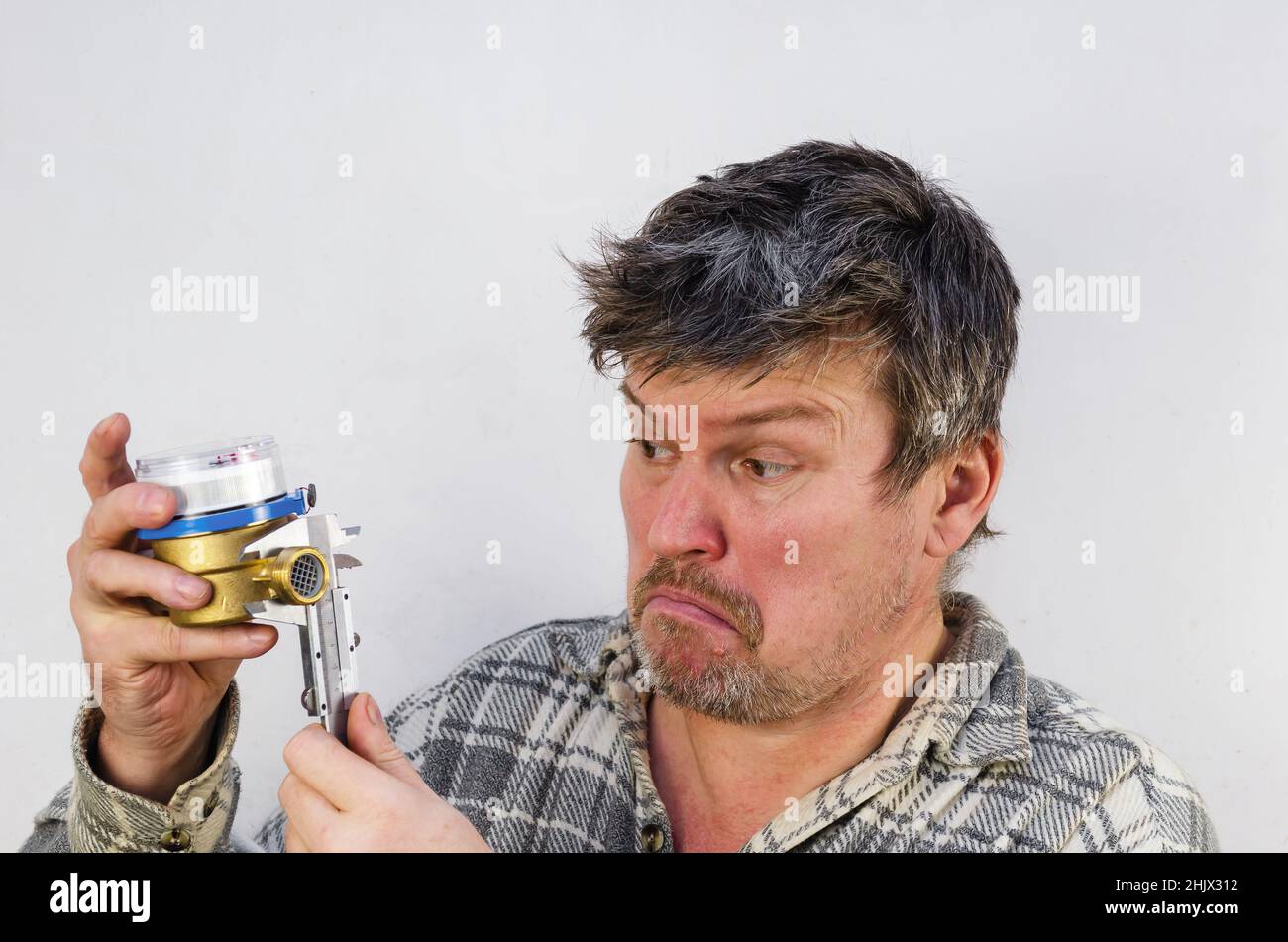 Il volto sorpreso di un uomo che misura un contatore dell'acqua con un calibro. L'uomo tiene in mano il contatore dell'acqua e il misuratore. Un uomo con aggrovigliato, dis Foto Stock