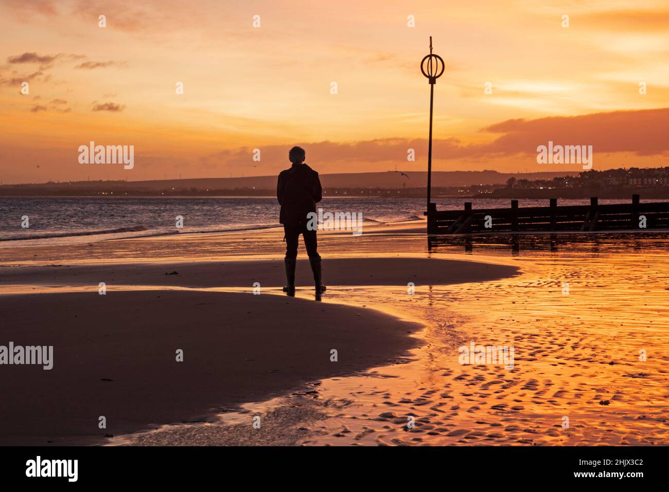 Portobello, Edimburgo, Scozia, Regno Unito. 1st febbraio 2022. Mite e ventose iniziano a febbraio poco prima dell'alba, quelle sulla spiaggia sabbiosa dal Firth of Forth salutate da un cielo colorato. Temperatura 9 gradi centigradi con vento ovest a 35km/h, raffiche potenziali di 72km/h. Nella foto: A volte il fotografo deve attivare il timer automatico della fotocamera e entrare nella scena. Foto Stock