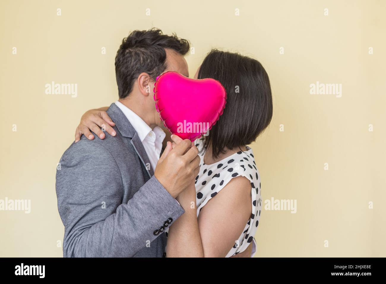 Vista laterale di coppia inconfondibile elegante baciare e nascondere le facce dietro il pallone a forma di cuore, mentre esprime l'amore il San Valentino beige Foto Stock