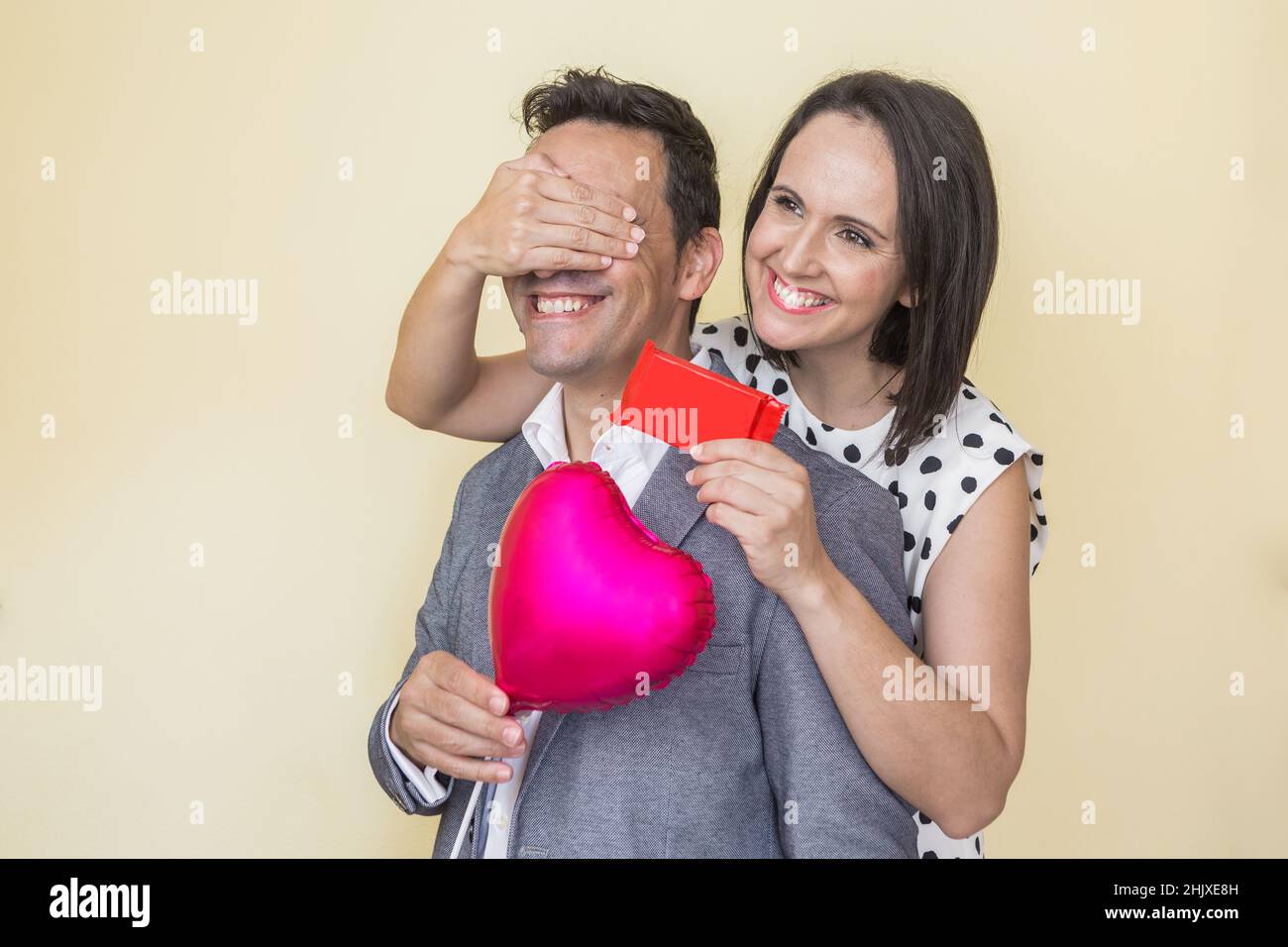 Allegra donna ispanica che copre gli occhi dell'uomo e gli dà presente mentre festeggia San Valentino su sfondo beige Foto Stock