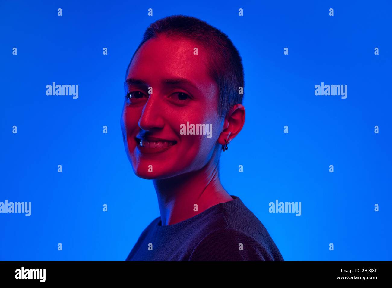 Ritratto di giovane donna sorridente, studente con capelli corti in maglione nero isolato su sfondo blu scuro in luce al neon. Concetto di emozioni Foto Stock