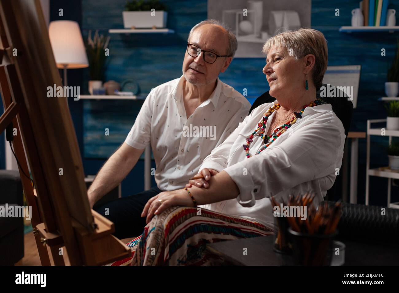 Coppia anziana che guarda il disegno seduto di fronte al cavalletto ammirando la creazione in casa studio d'arte. Vecchi artisti creativi pensionati che si sentono orgogliosi di opere d'arte in officina. Foto Stock