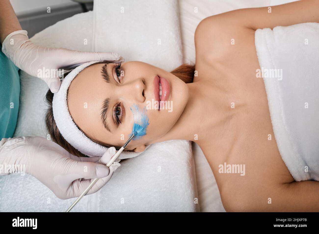 Donna durante l'applicazione della maschera cosmetica per la cura del viso con estetista sdraiato nel salone di cosmetologia, vista dall'alto. cura della pelle del viso con cosmetici mas Foto Stock