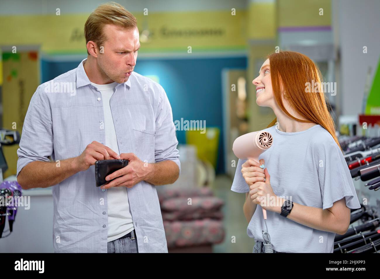 Cute Redhead femmina chiedere a marito da asciugacapelli, elemosina. L'uomo non ha soldi, tenendo la borsetta nelle mani, al negozio di elettrodomestici. Adorabile Woma adulto Foto Stock
