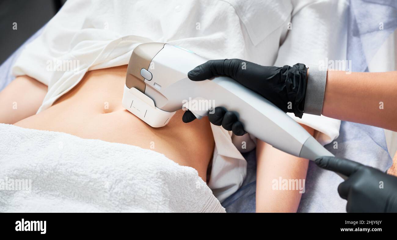 Primo piano medico cosmetologo utilizzando il dispositivo a ultrasuoni durante l'esecuzione della procedura di sollevamento su donna addome. Paziente femmina che riceve la procedura di irrigidimento e contornazione del corpo in clinica di cosmetologia. Foto Stock