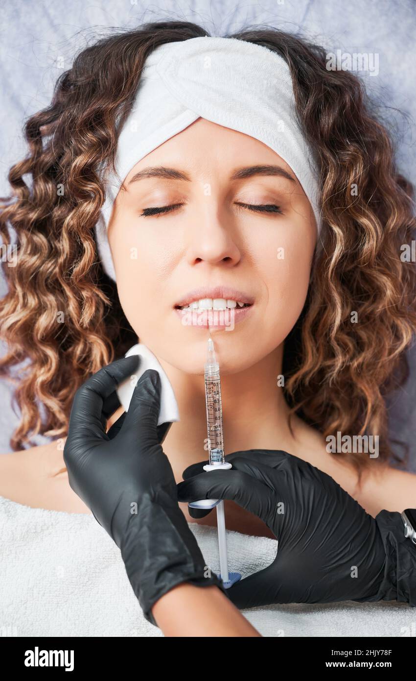 Primo piano ritratto di attraente giovane donna con capelli ricci sdraiati ed estetista facendo iniezione in labbro con preparazione speciale. Concetto di procedura di processo aumento del labbro in salone professionale. Foto Stock