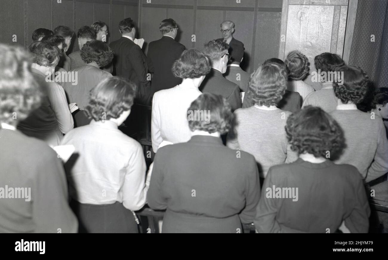 1950s, storica, congregazione cristiana.. uomini e donne che si levano in piedi insieme in una piccola stanza, leggendo dai bibbi. Un pastore maschile in un vestito e cravatta, si trova all'estremità della stanza, di fronte al suo pubblico, USA. Foto Stock