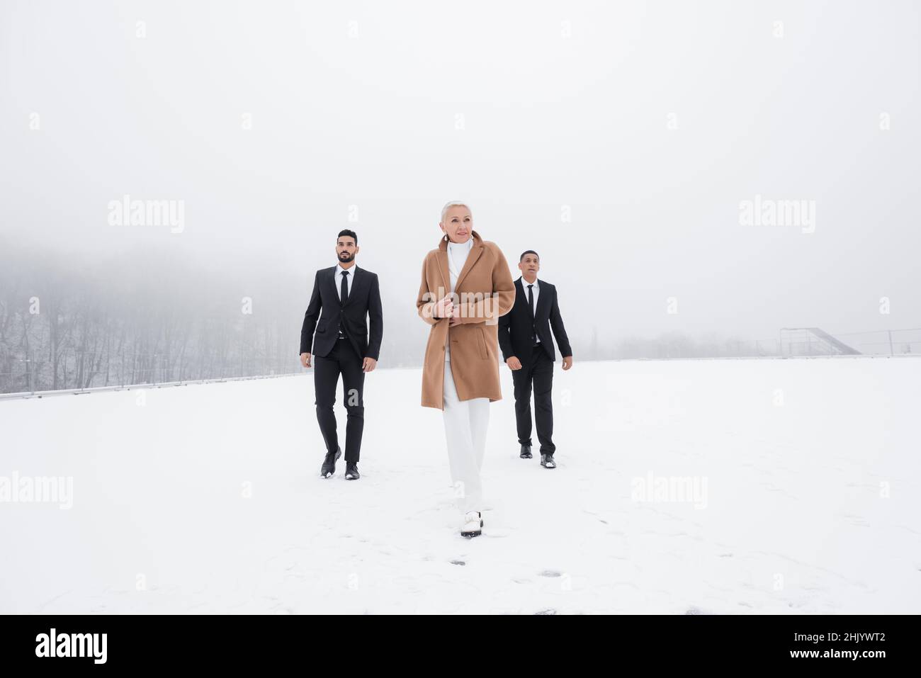 vista a tutta lunghezza dell'elegante uomo d'affari anziano che cammina attraverso il campo invernale con le guardie del corpo interrazziali Foto Stock