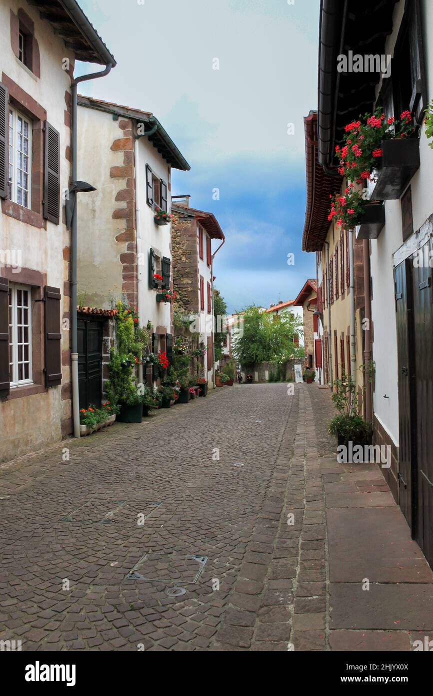 Le case bianche con pietre di taglia, gerani rossi sono appesi sulle facciate, la strada scende verso altre case, è il Paese basco francese Foto Stock