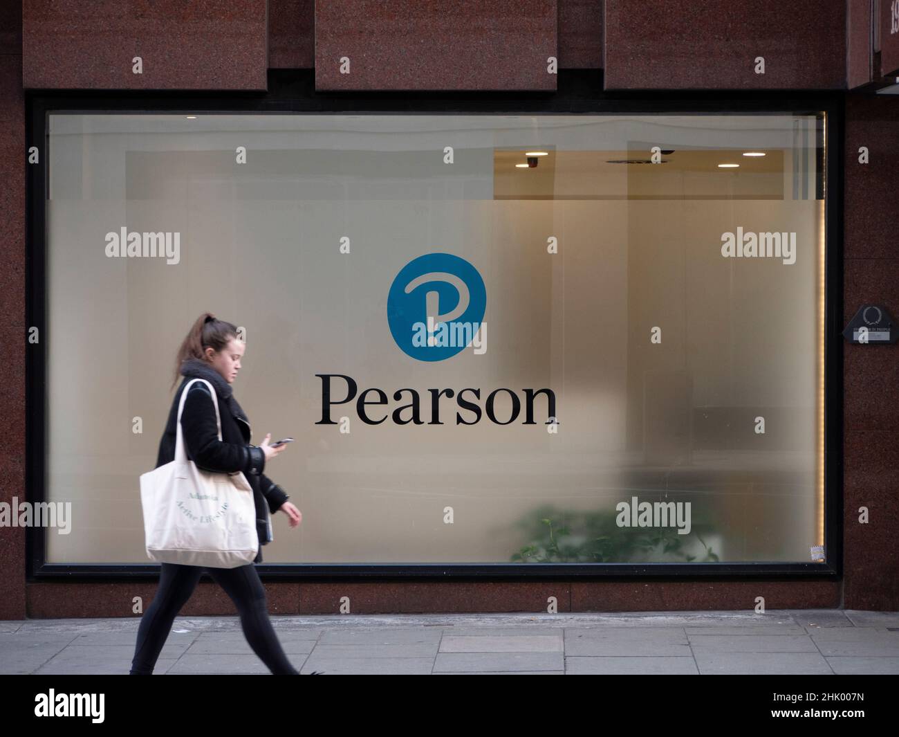 Donna che guarda il dispositivo mobile del telefono mentre cammina oltre la sede centrale dell'azienda di formazione Pearson nel centro di Londra, Pearson plc è un'azienda britannica di editoria e formazione con uffici a Londra. Foto Stock