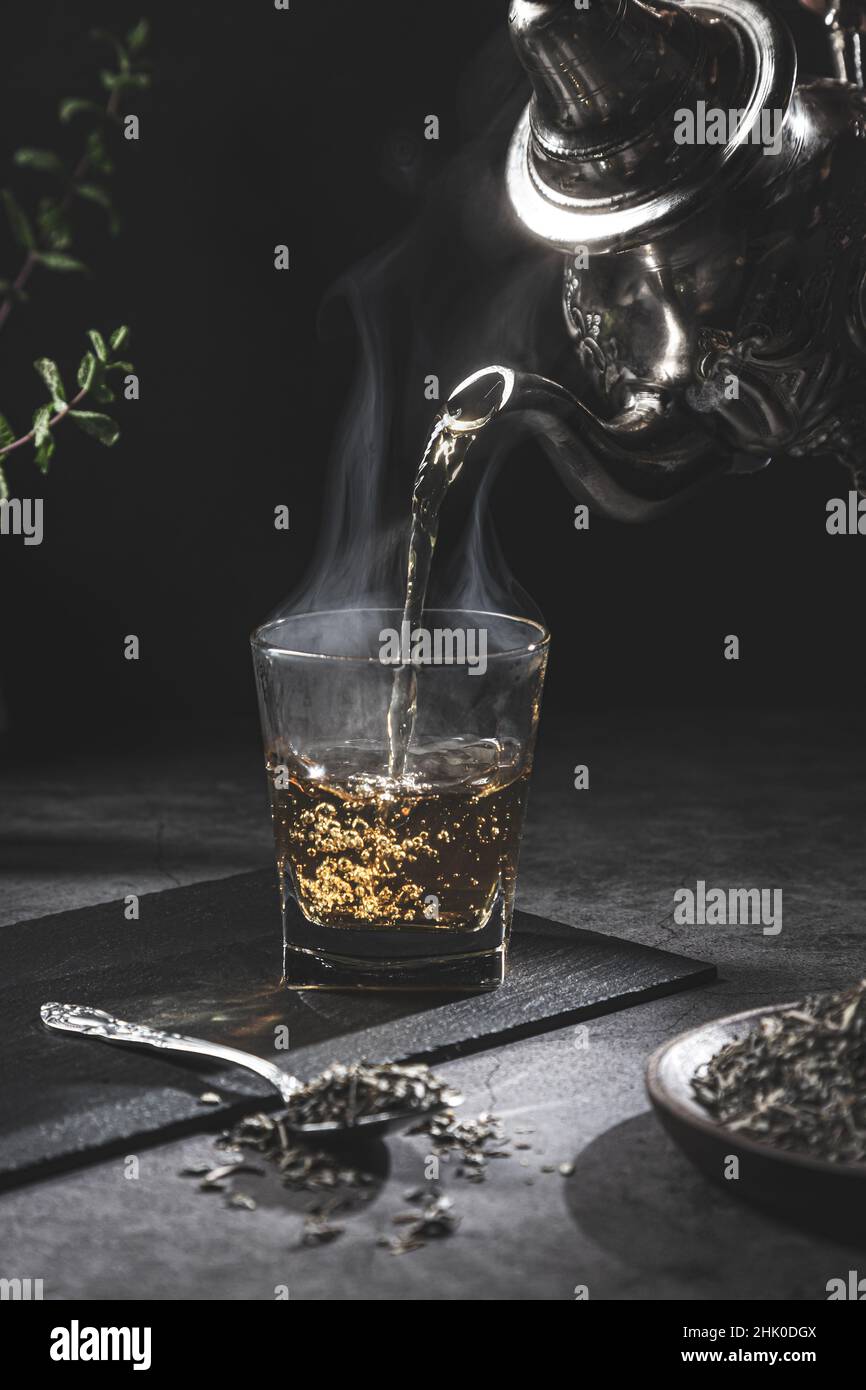 Tradizionale teiera marocchina che versa il tè in un bicchiere fumante con tè naturale e menta su sfondo scuro. Tradizionale concetto di tè marocchino. Foto Stock