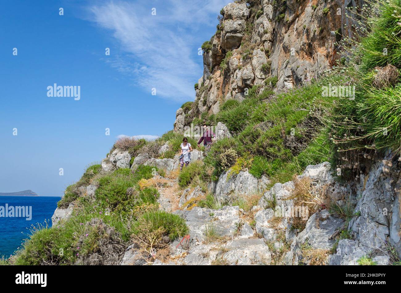 La gente cammina e si arrampica attraverso l'ambiente naturale con cespugli e ripide formazioni rocciose a Monemvasia, Grecia. Scena estiva con cielo blu e mare Foto Stock