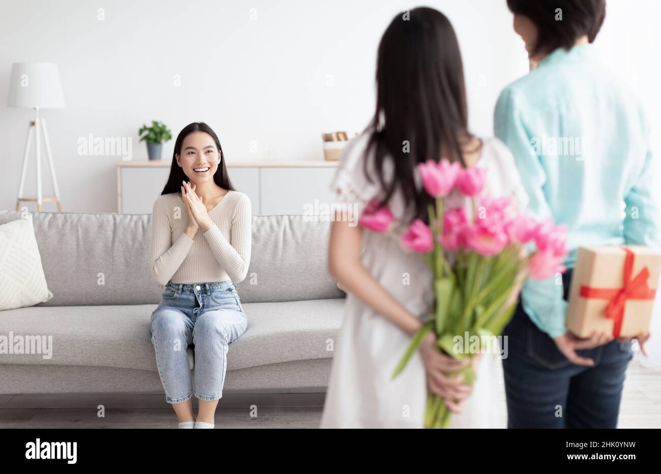 Piccola ragazza asiatica con il suo regalo e i fiori per la giovane donna dietro le spalle a casa. Vacanza in famiglia e festeggiamenti concetto Foto Stock