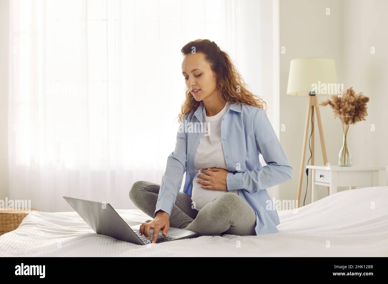 Donna incinta seduta a letto in camera da letto e utilizzando un computer portatile alla ricerca di informazioni sulla gravidanza. Foto Stock