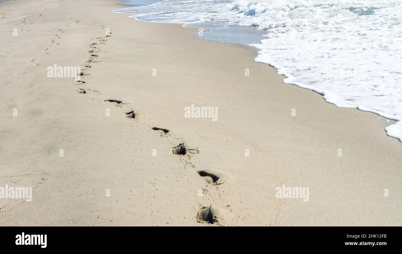 Una catena solitaria di impronte di una persona sulla sabbia sulla riva del mare accanto all'onda ondulata. Passeggiata lungo il mare. Il concetto di solitudine Foto Stock