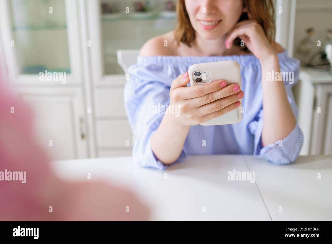 Felice donna brunette in elegante blusa blu guarda nel telefono cellulare sorridente e seduto al tavolo bianco in camera luminosa a casa vista ravvicinata Foto Stock