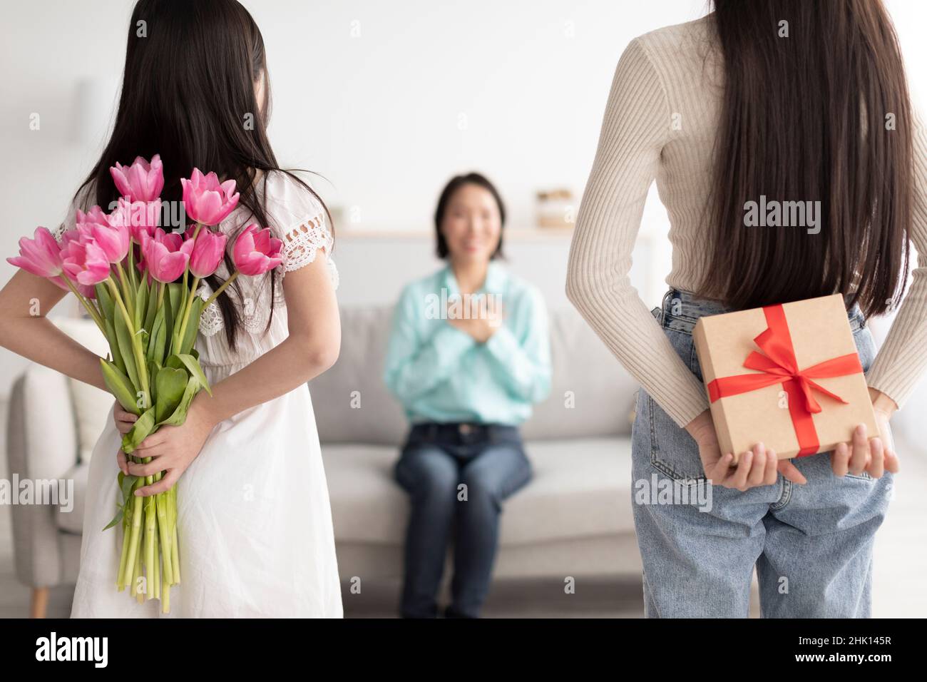 La giovane donna e la figlia nascondono il dono e i tulipani dietro le spalle, congratulandosi con la donna matura con le vacanze primaverili Foto Stock