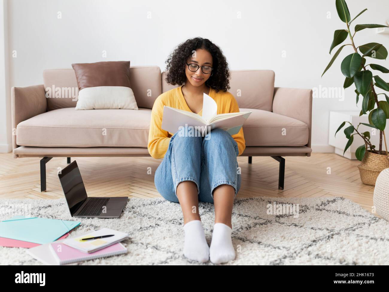 Bella giovane donna nera libro di lettura, seduta sul pavimento vicino al PC portatile e materiali di studio a casa Foto Stock