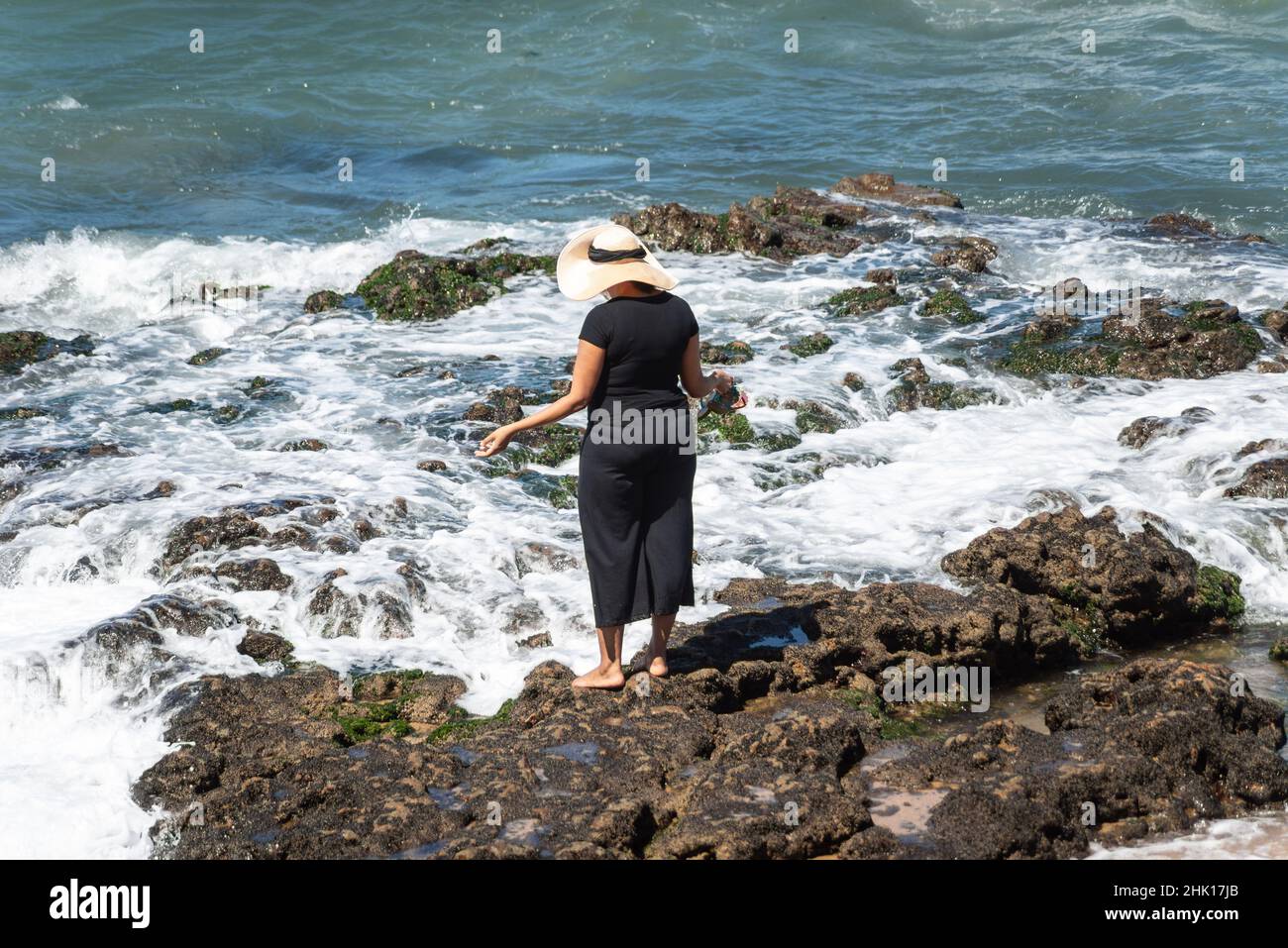 La donna in abiti neri e cappello in piedi sulle rocce della spiaggia di Farol da barra a Salvador, Bahia, Brasile Foto Stock