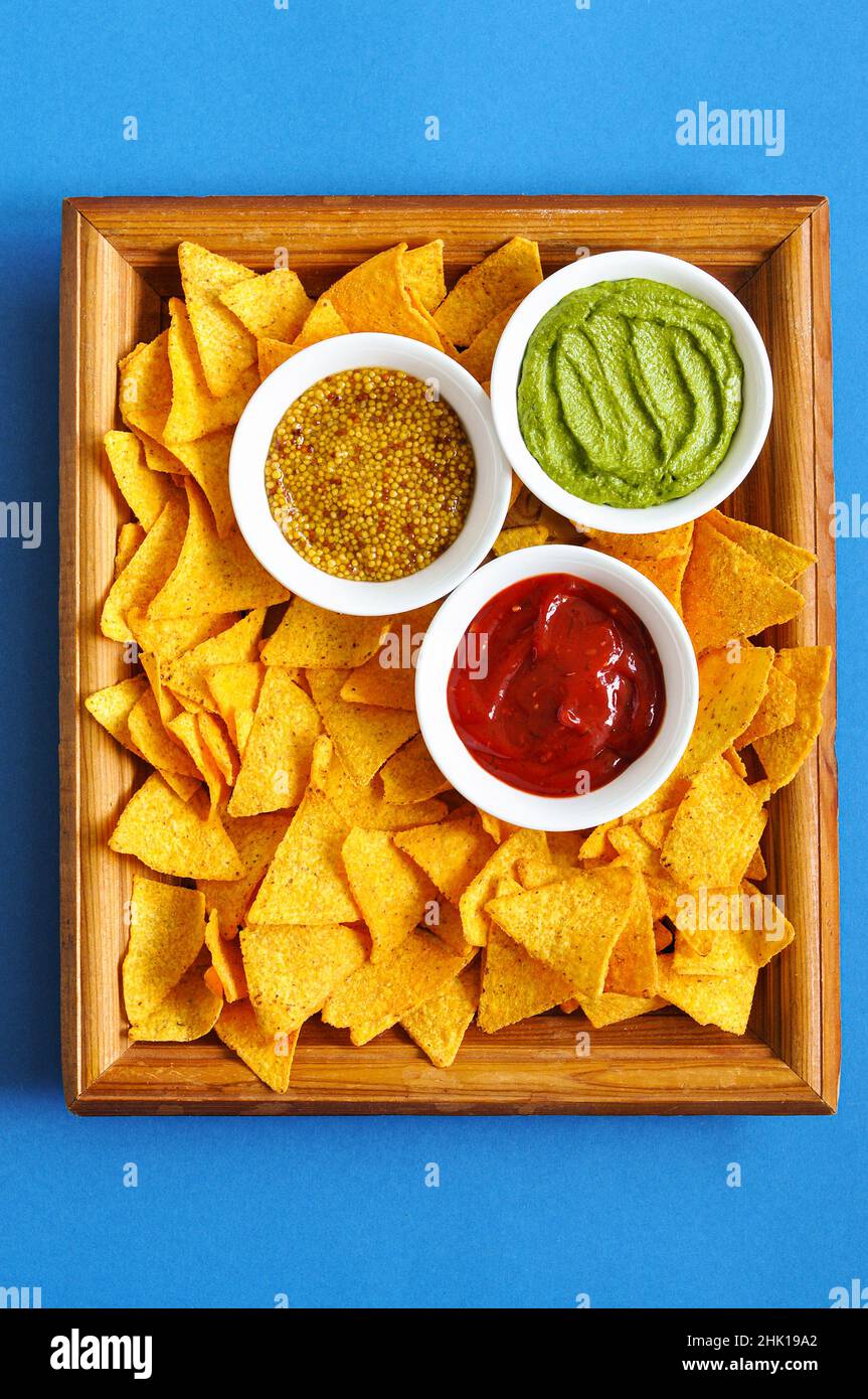 Mucchio di tortilla di mais con mostarda francese, guacamole e ketchup servito su un asse di legno su sfondo blu. Parte superiore ivew. Foto Stock